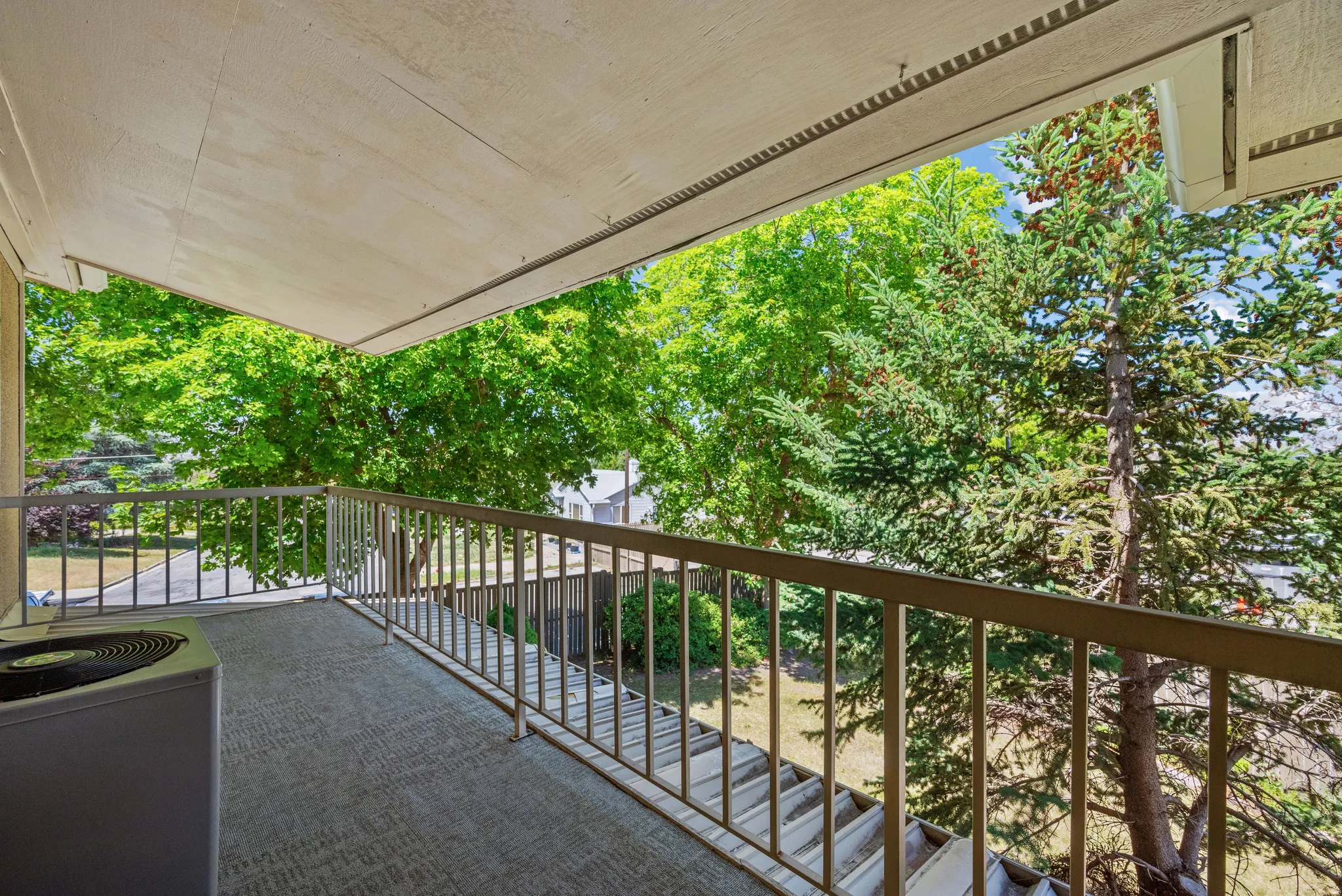 Private covered balcony with a new outdoor carpet surface and metal railing, surrounded by mature trees