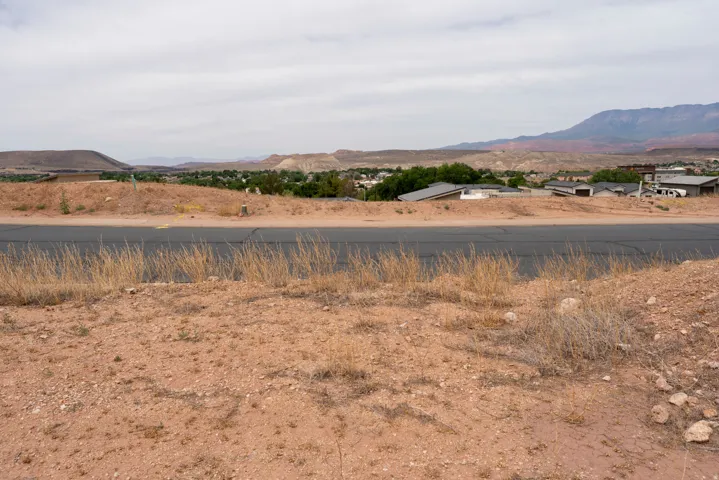 Elevated desert lot featuring a paved road frontage, distant mountain backdrop, and scattered residential structures