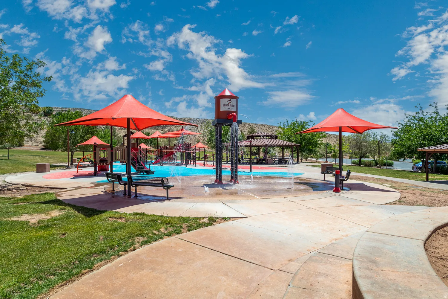 Community splash pad featuring multiple water features, shaded seating areas, and a nearby playground structure