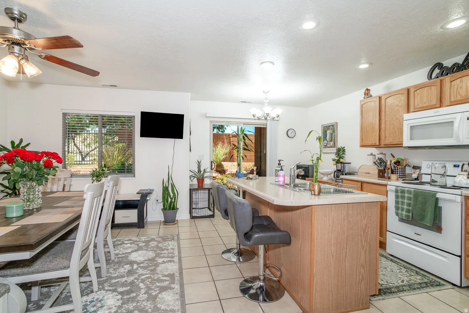 Open-concept kitchen and dining area featuring tile flooring, light wood-finish cabinetry, a kitchen island with a built-in sink, and a dining area with a ceiling fan