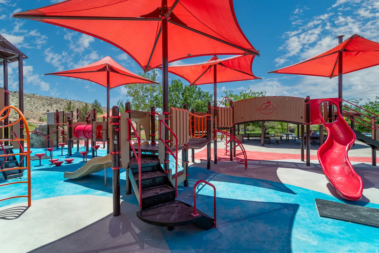 Expansive playground featuring multiple play structures, red shade canopies, and a soft, multi-colored ground surface