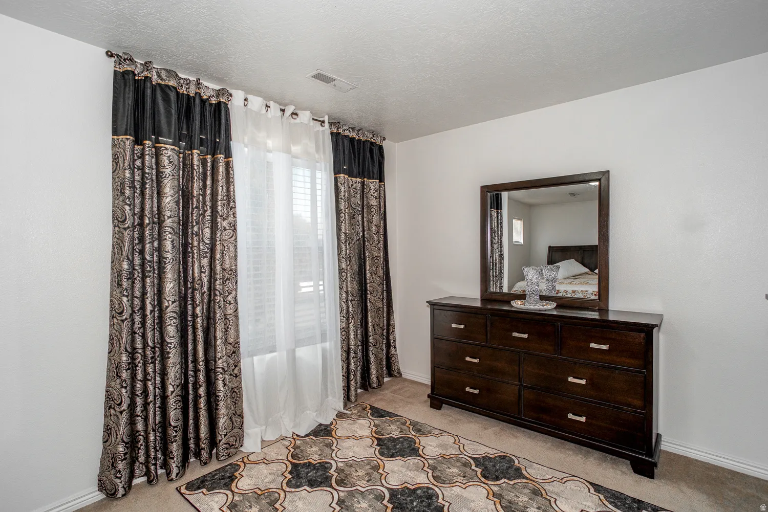 Bright room featuring a window with white blinds and layered drapery, neutral carpet flooring, and a ceiling vent