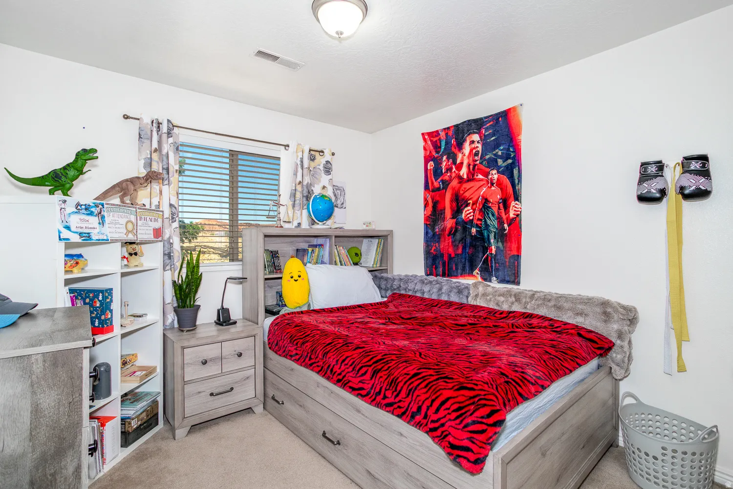 Carpeted bedroom featuring a window with horizontal blinds, built-in shelving, a wood-finish nightstand with drawers, and a ceiling-mounted light fixture