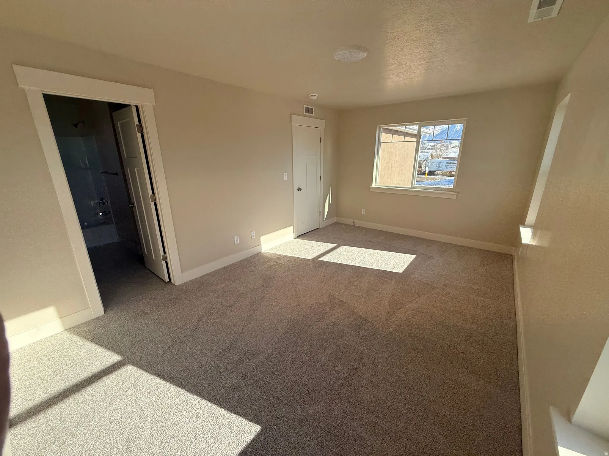 Carpeted room with neutral-toned walls, featuring a window and an entry to a bathroom