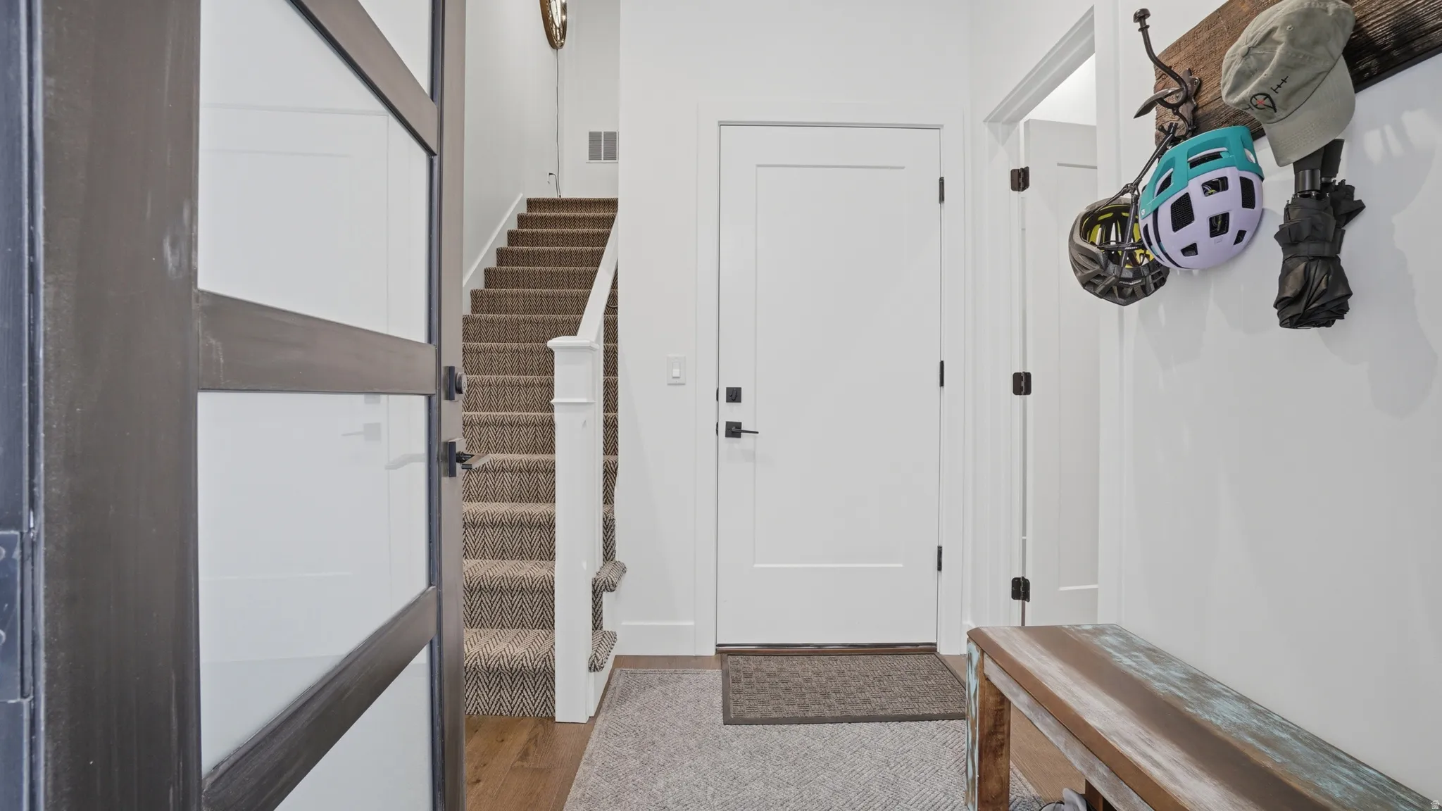 Entryway featuring a white solid-core door with black hardware, wood-finish flooring, a carpeted staircase with white risers, and a white baluster