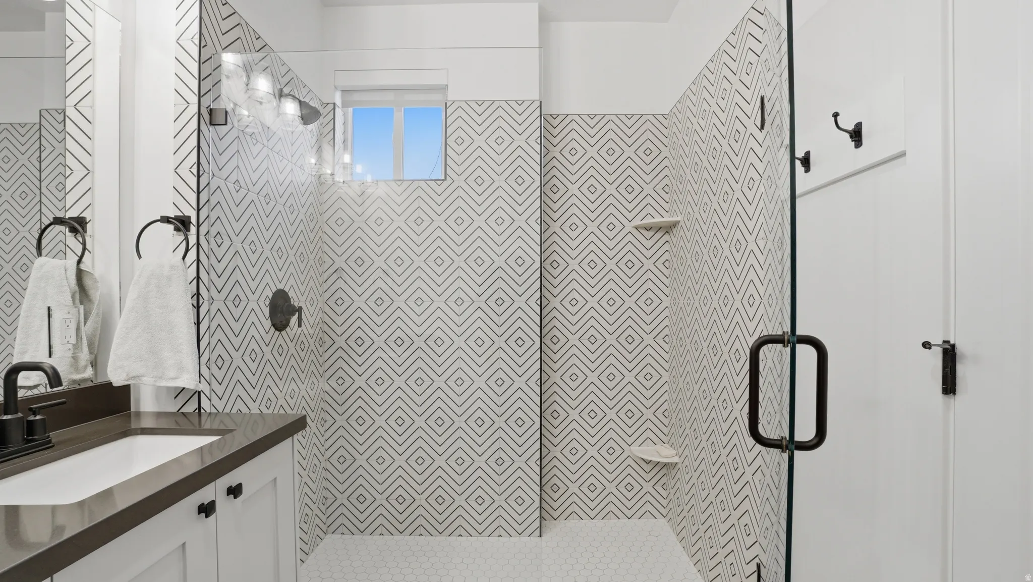 Contemporary bathroom featuring a walk-in shower with geometric patterned tile and a frameless glass enclosure, a window, hexagonal mosaic floor tile, a single vanity with a rectangular sink, and dark-finish hardware