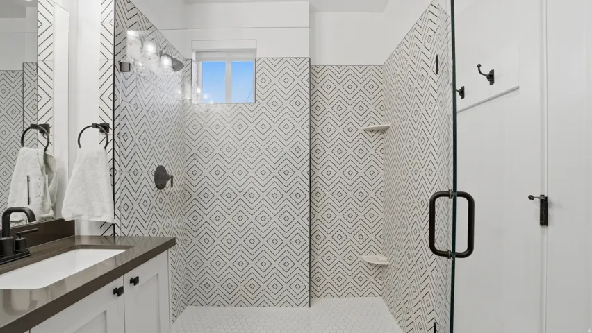 Contemporary bathroom featuring a walk-in shower with geometric patterned tile and a frameless glass enclosure, a window, hexagonal mosaic floor tile, a single vanity with a rectangular sink, and dark-finish hardware
