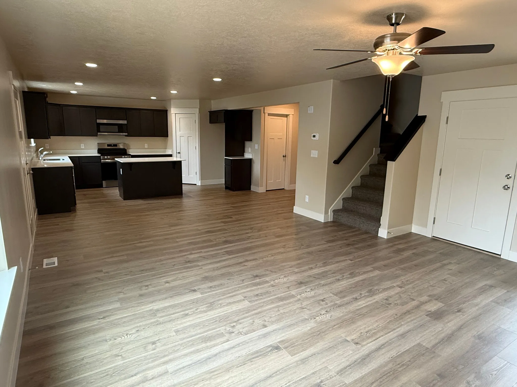 Expansive main living area featuring wood-finish flooring, a contemporary kitchen with dark cabinetry and light countertops, stainless steel appliances, recessed lighting, and a ceiling fan