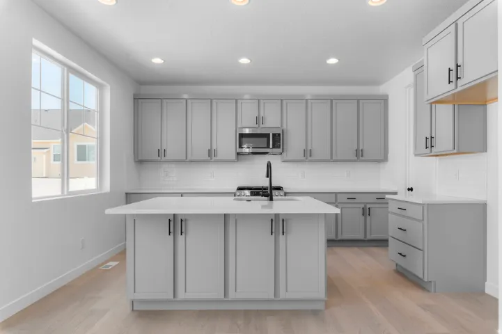 Kitchen featuring light wood-finish flooring, recessed lighting, a center island with sink and matte black faucet, gray cabinetry with black hardware, and white subway tile backsplash