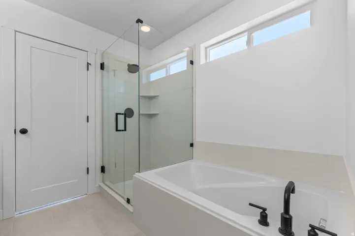 Soaking tub with matte black fixtures, separate glass-enclosed shower with built-in shelving, and transom windows
