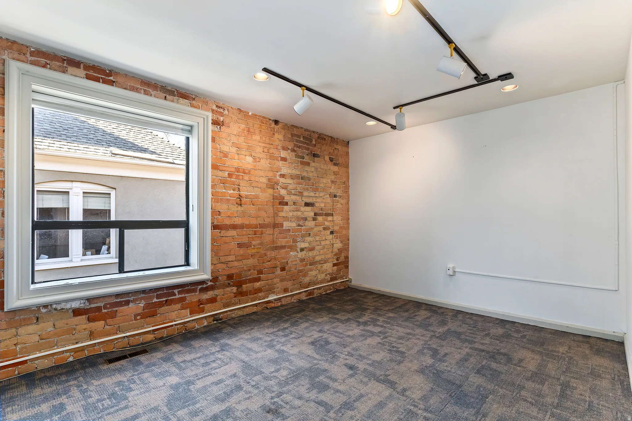 Exposed brick wall with a large window and white trim, featuring track lighting and carpet flooring