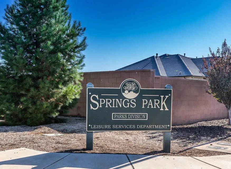Springs Park sign with "Parks Division" and "Leisure Services Department" visible, featuring a mature evergreen tree, a low-lying deciduous tree, a block wall structure, and a residential roofline in the background