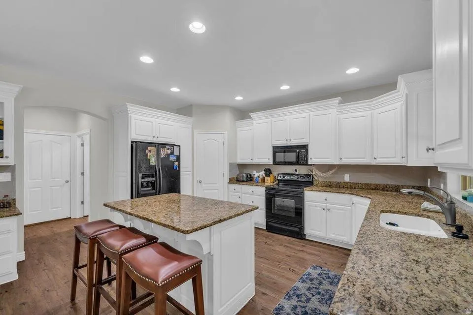 Kitchen featuring an island with stone countertop, white cabinetry, integrated black appliances, wood-finish flooring, and recessed lighting