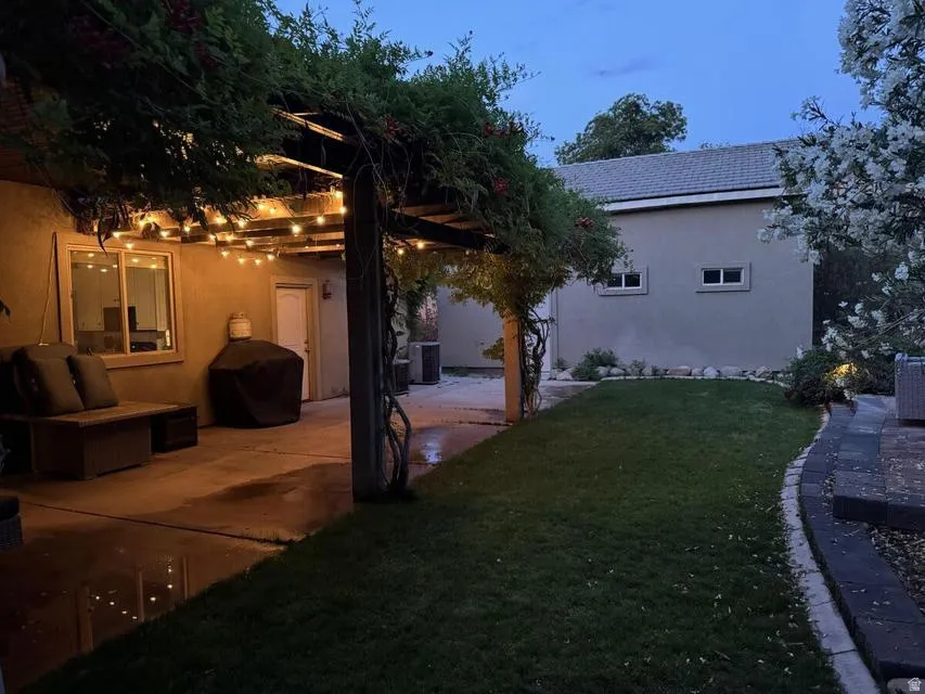 Spacious patio area featuring an overhead pergola with string lighting, a manicured lawn, and a stone-edged flower bed