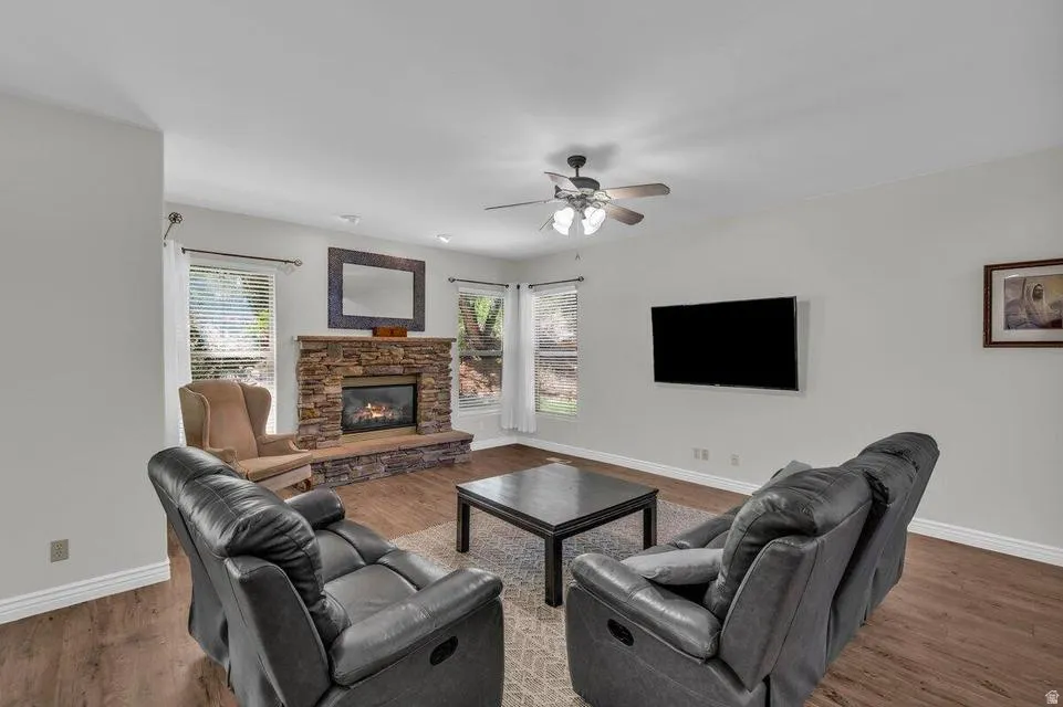 Living area featuring wood-finish flooring, a stone-faced fireplace with a hearth, multiple windows, a ceiling fan with integrated lighting, and white baseboards