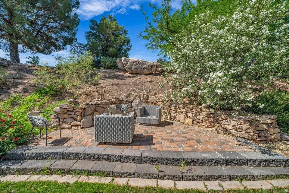 Brick paver patio with a stone retaining wall and tiered block steps, surrounded by mature trees and flowering shrubs