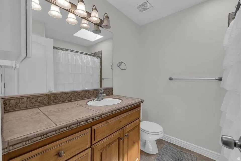 Bathroom featuring a wood-finish vanity with an integrated sink and tile countertop, large vanity mirror, and skylight