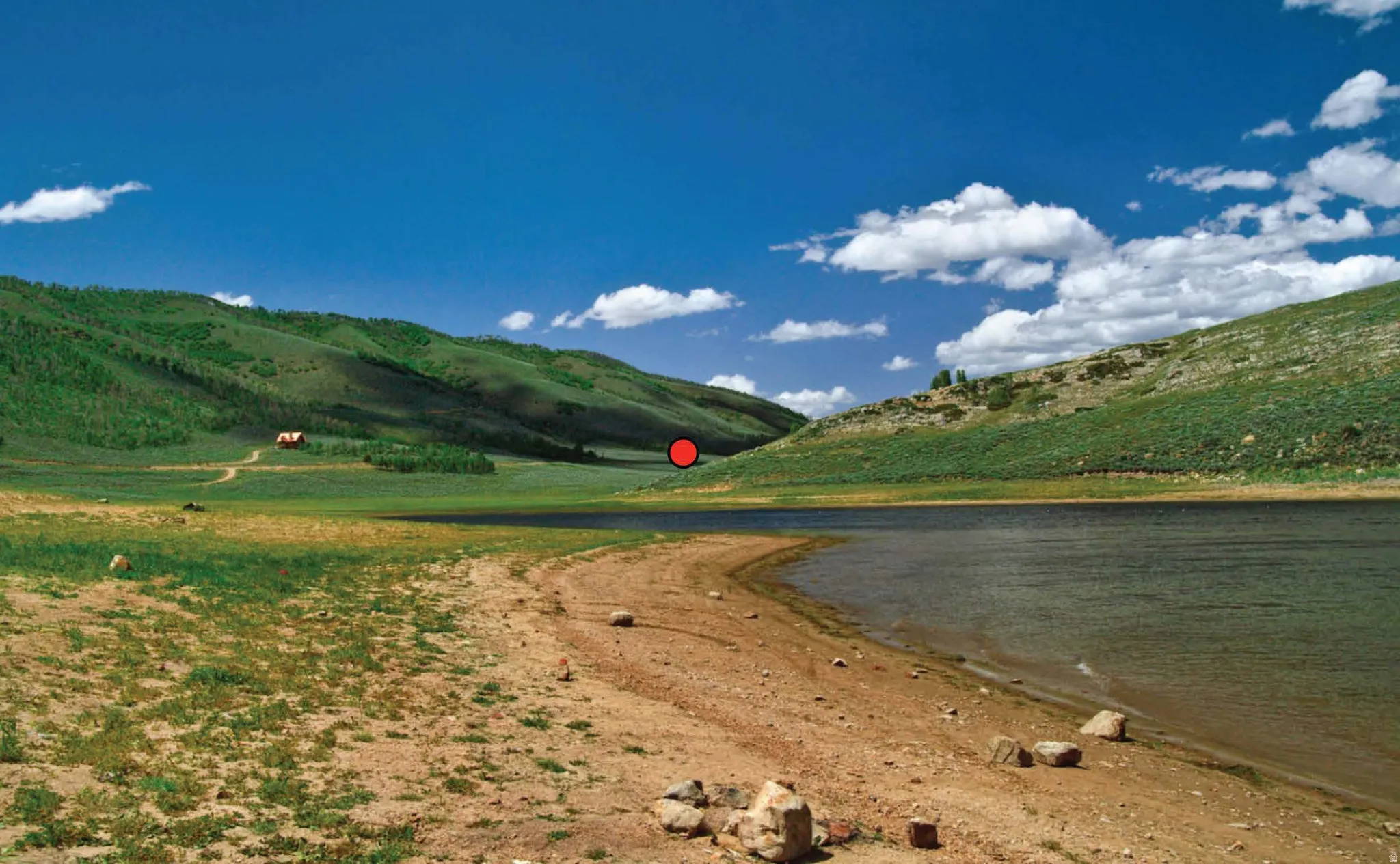 Lakefront property featuring a natural shoreline, rolling green hills, a distant building, and a clear blue sky
