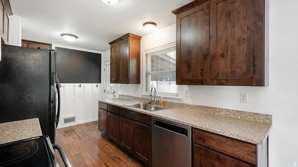 Kitchen featuring wood-finish flooring, dark wood cabinetry, light speckled countertops, a stainless steel double sink, and a black appliance package