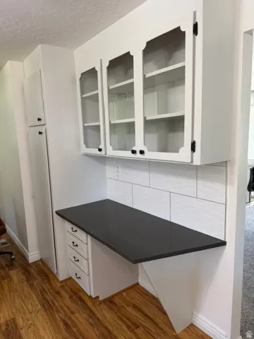 Built-in desk featuring a dark countertop, white shaker-style drawers, and overhead glass-front cabinetry