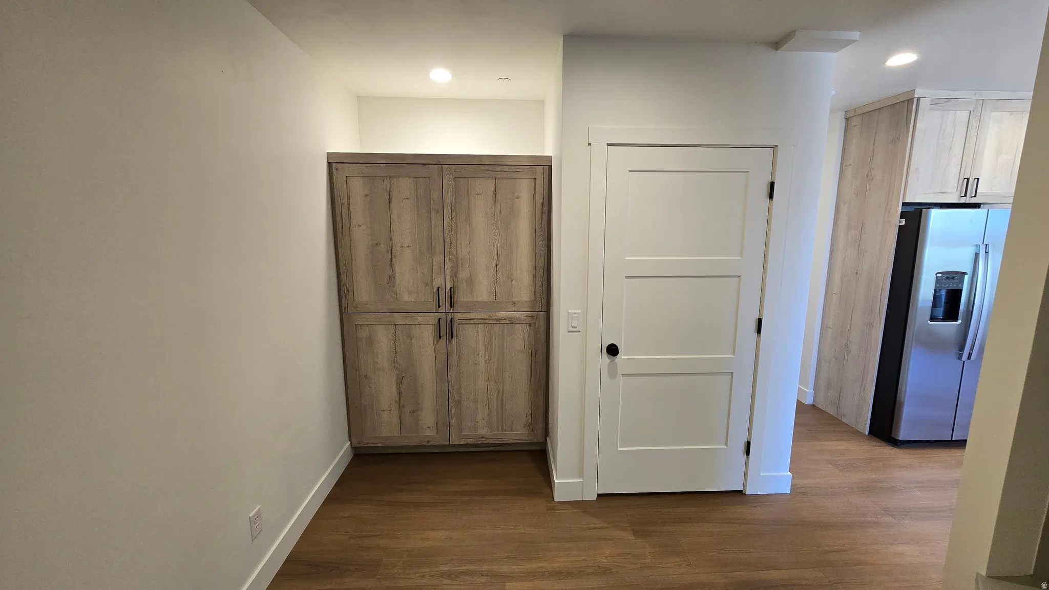 Built-in wood-finish cabinetry, recessed ceiling lighting, wood-finish flooring, white paneled interior door with matte black hardware, and stainless steel refrigerator
