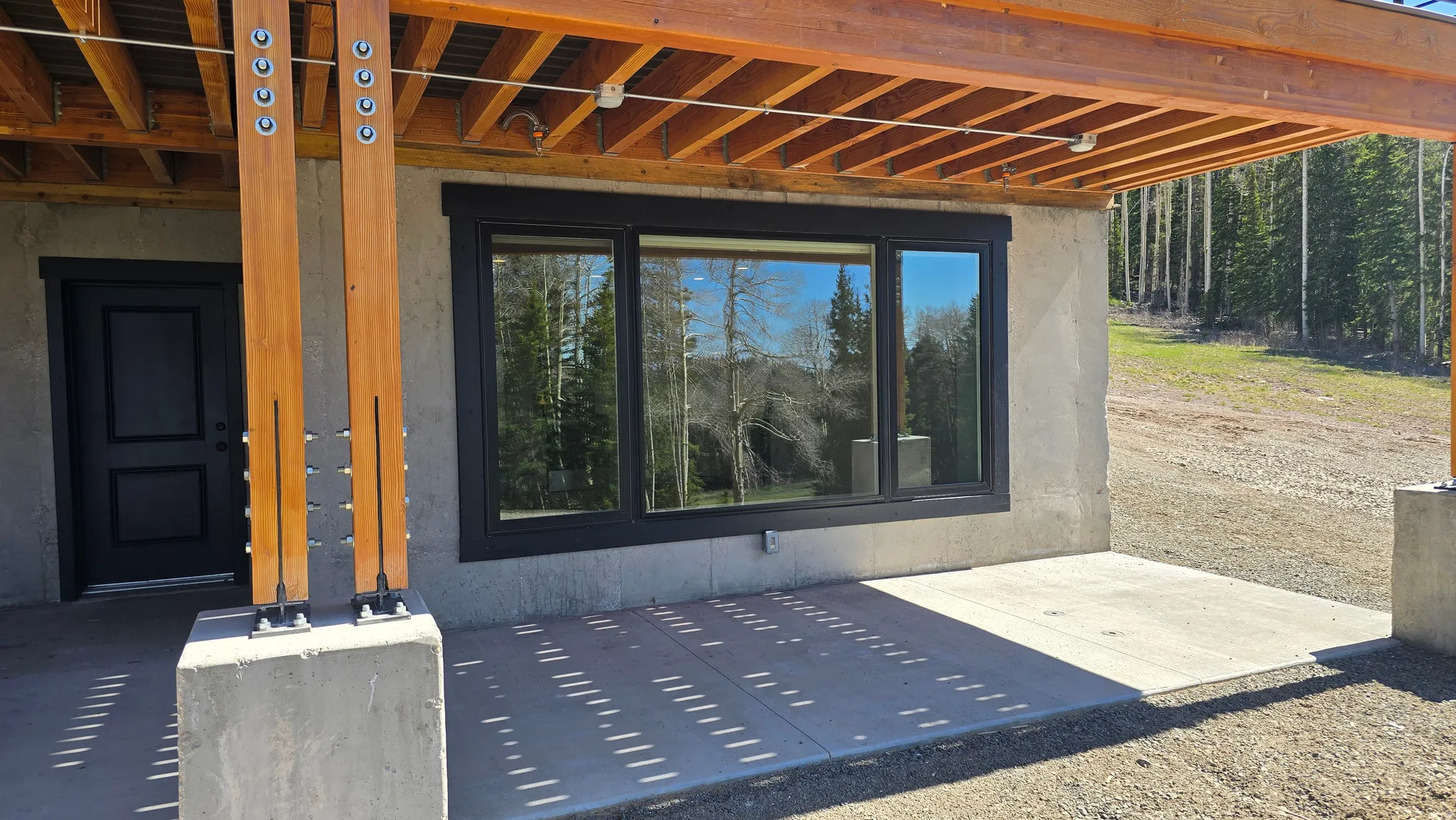 Covered concrete patio with exposed timber beams, large picture windows, and a solid black entry door