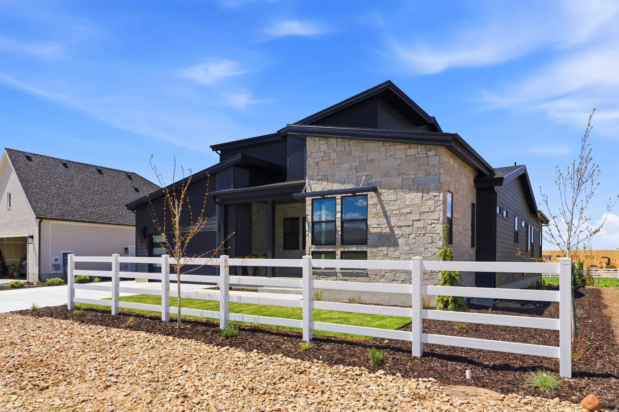 Modern architectural design featuring stone and dark siding, multiple large windows, a covered entry, and a white fence