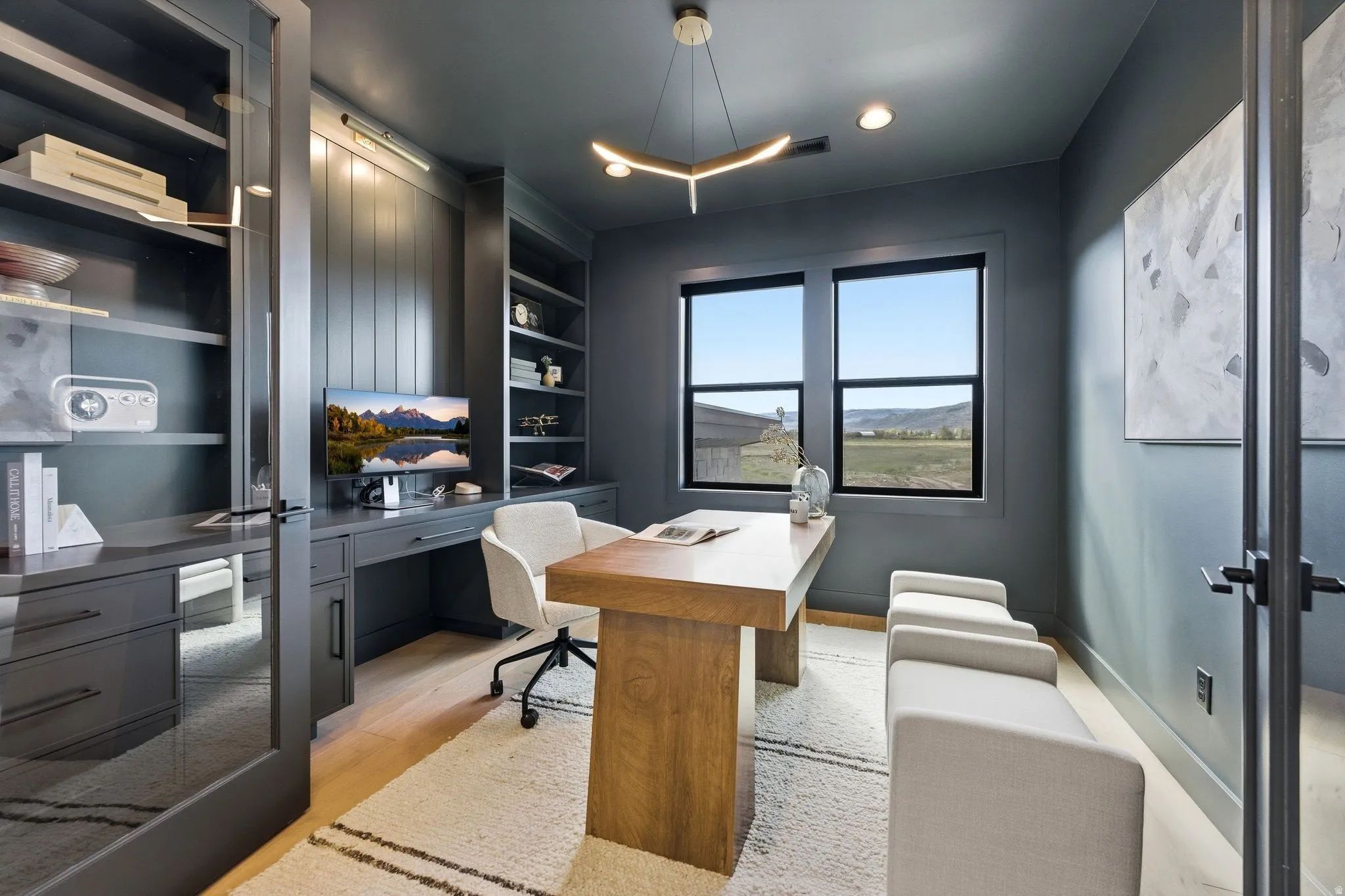 Dedicated office space featuring two large windows, built-in shelving, a full-wall desk unit, and light wood-finish flooring