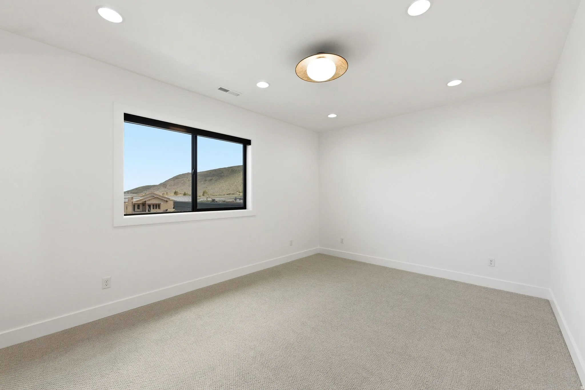 Light-filled room featuring a broad window with black framing, recessed ceiling lighting, a central flush-mount fixture, high baseboards, and light-toned carpeting