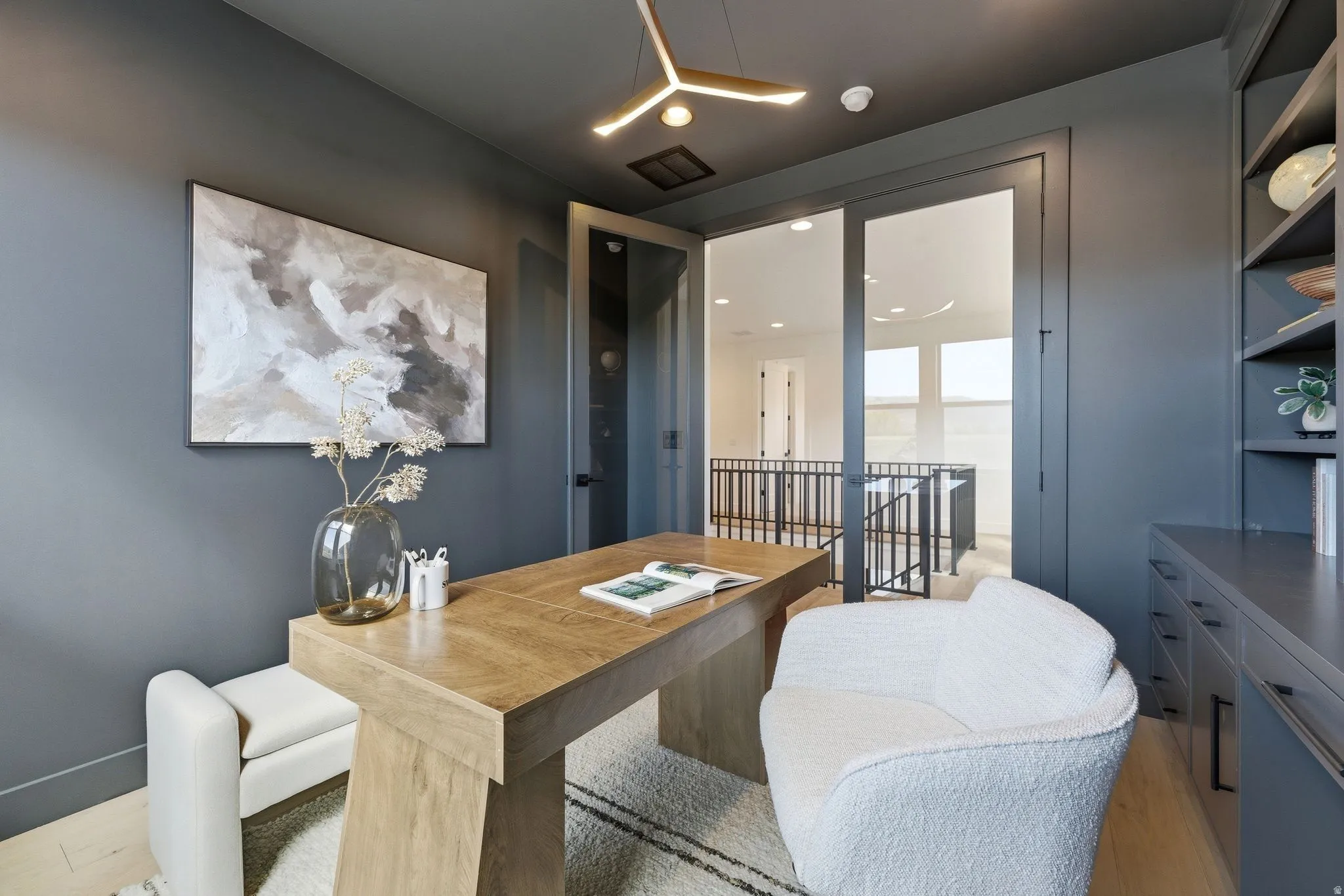 Dedicated office space featuring a built-in shelving unit, custom cabinetry, modern lighting fixture, wood-finish flooring, and glass-paneled double doors