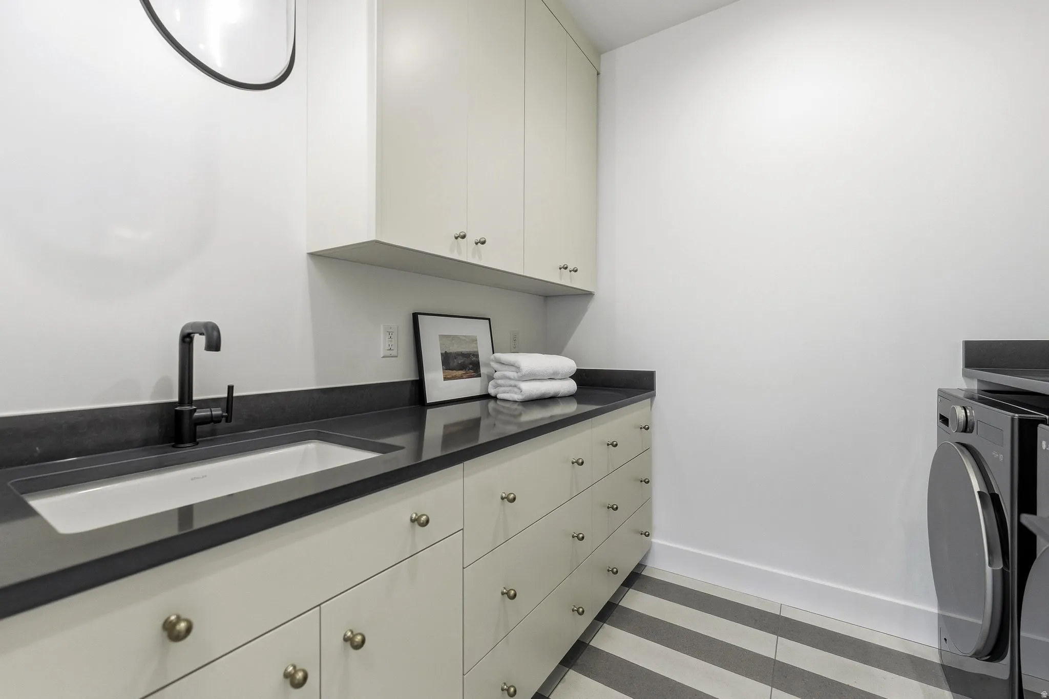 Utility room featuring light-colored cabinetry with brass hardware, a dark countertop, an undermount sink with a matte black faucet, and striped tile flooring