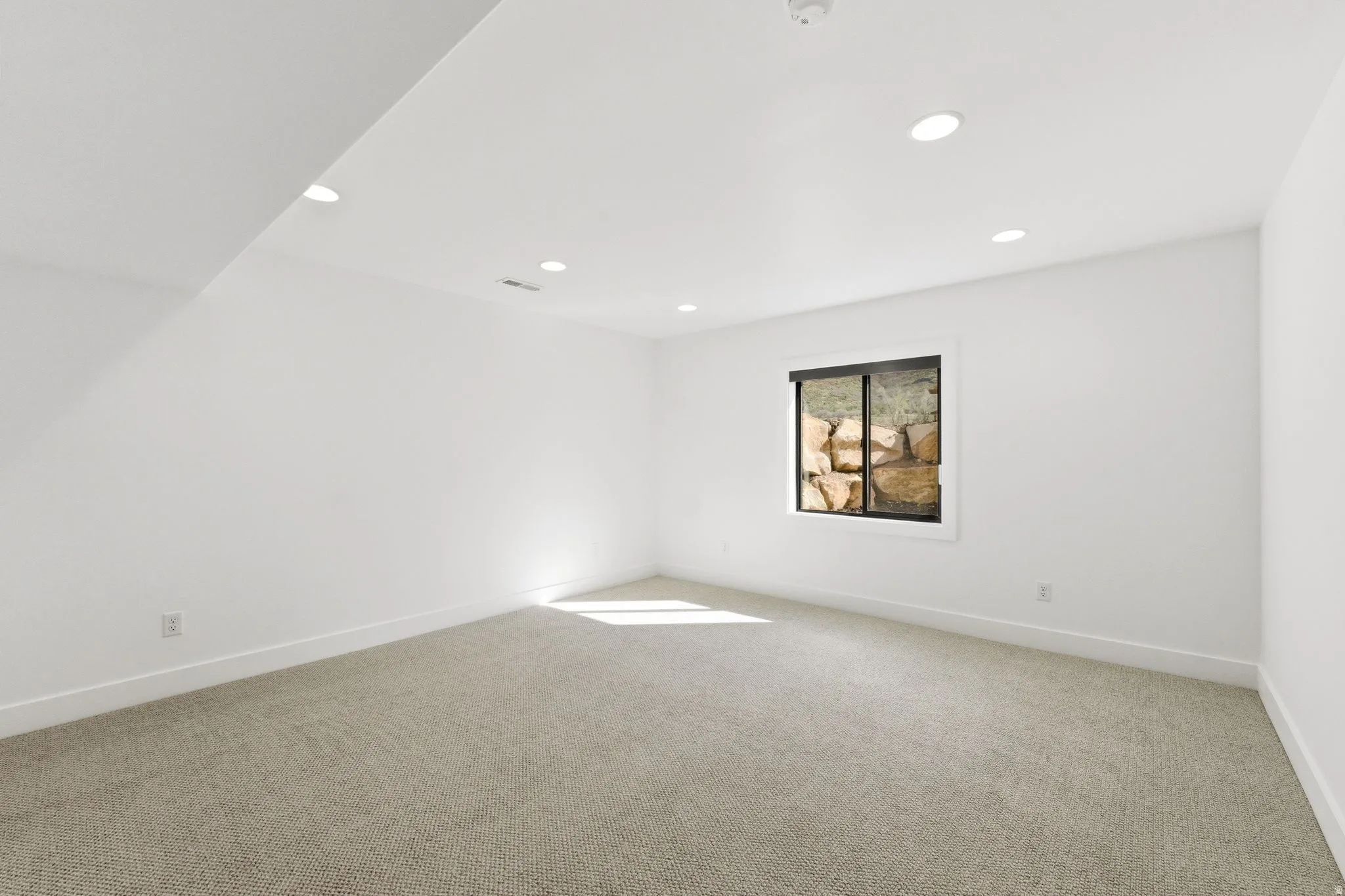 Spacious room with recessed lighting, light-toned carpeting, white walls, and a window featuring a natural rock backdrop