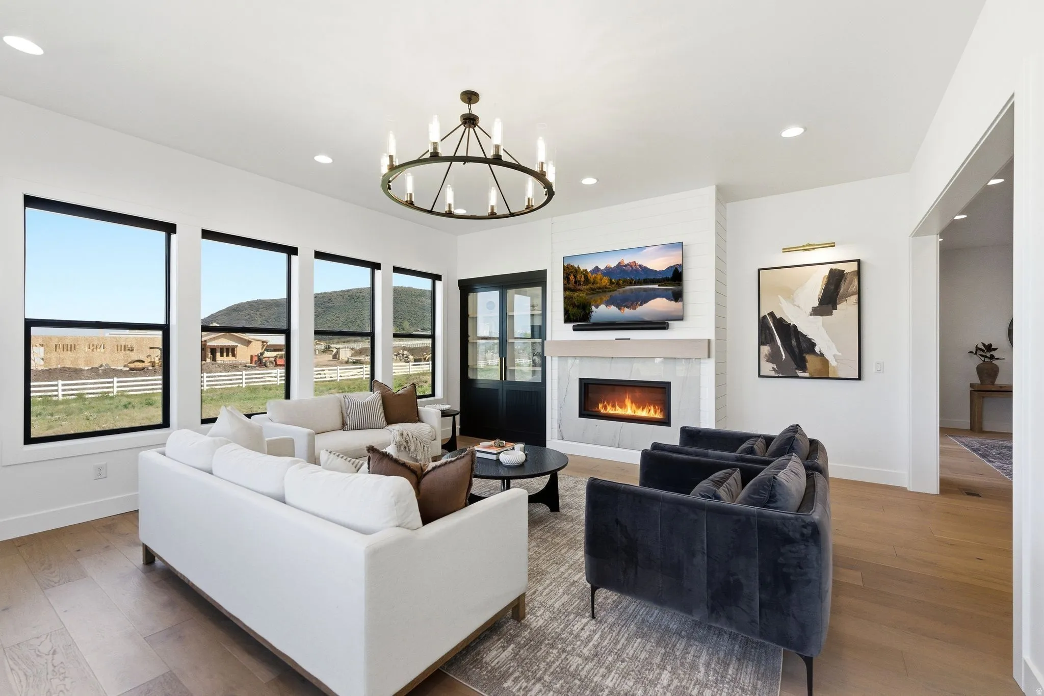Spacious living area featuring expansive windows, wood-finish flooring, a contemporary fireplace with a stone surround, recessed lighting, and an ornate chandelier