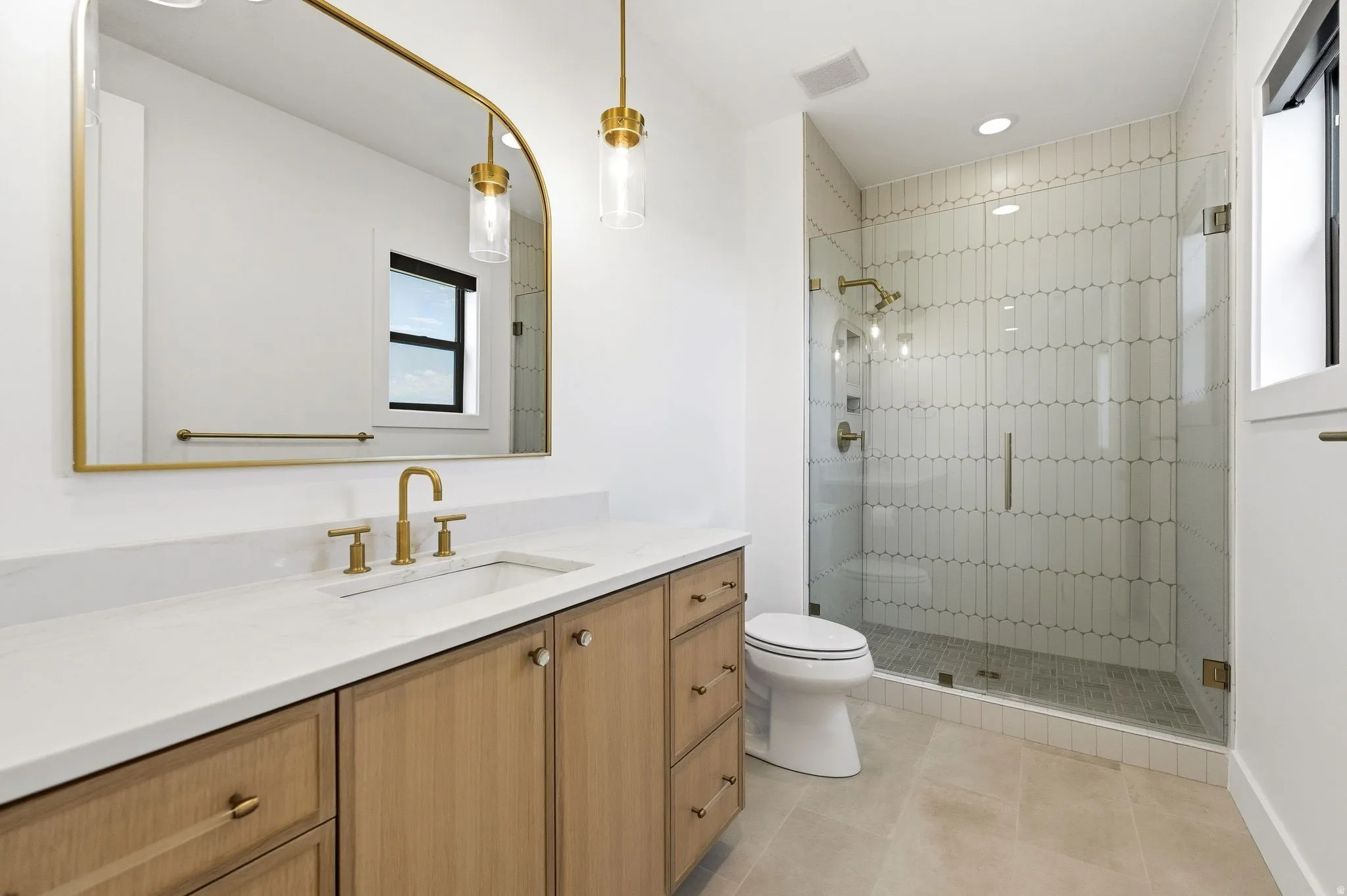 Bathroom featuring a light wood-finish vanity with a white countertop and undermount sink, brass-finish fixtures, an arched mirror, and pendant lighting