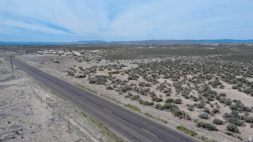 Expansive land parcel featuring a paved road with double yellow lines, utility poles along the roadside, and sparse vegetation, extending to distant mountains under a blue sky