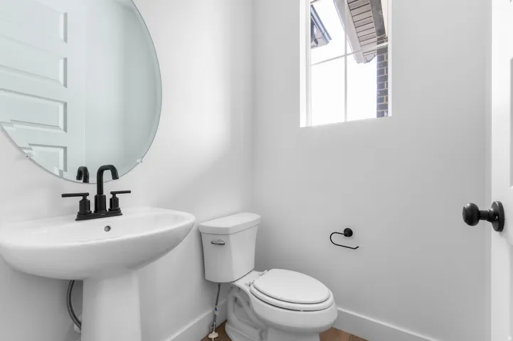 Pedestal sink with matte black faucet, wall-mounted circular mirror, window with exterior architectural detail, and white paneled door