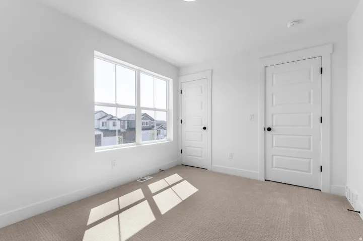 Bright corner bedroom featuring large multi-pane windows flooding the space with natural light, neutral wall finishes, neutral-toned carpeting, and two white six-panel doors providing flexible entry options
