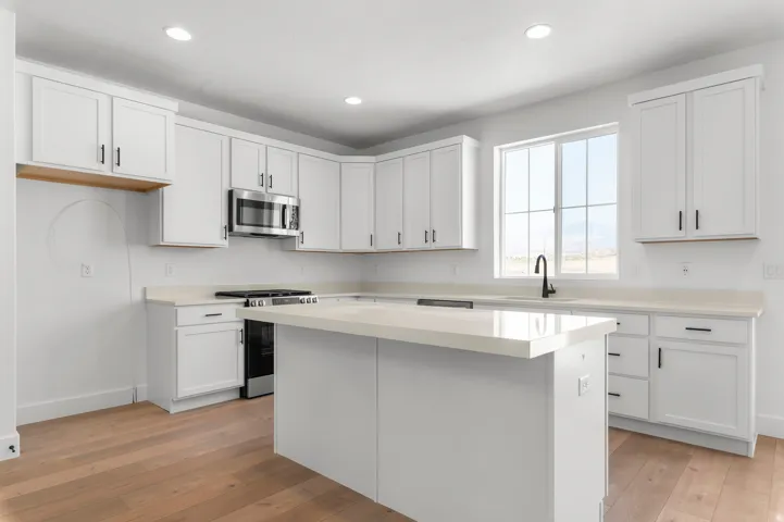 Modern kitchen featuring white shaker-style cabinetry, stainless steel appliances including a gas range and microwave, quartz countertops, a large center island with seating, recessed ceiling lighting, and wood-finish flooring throughout
