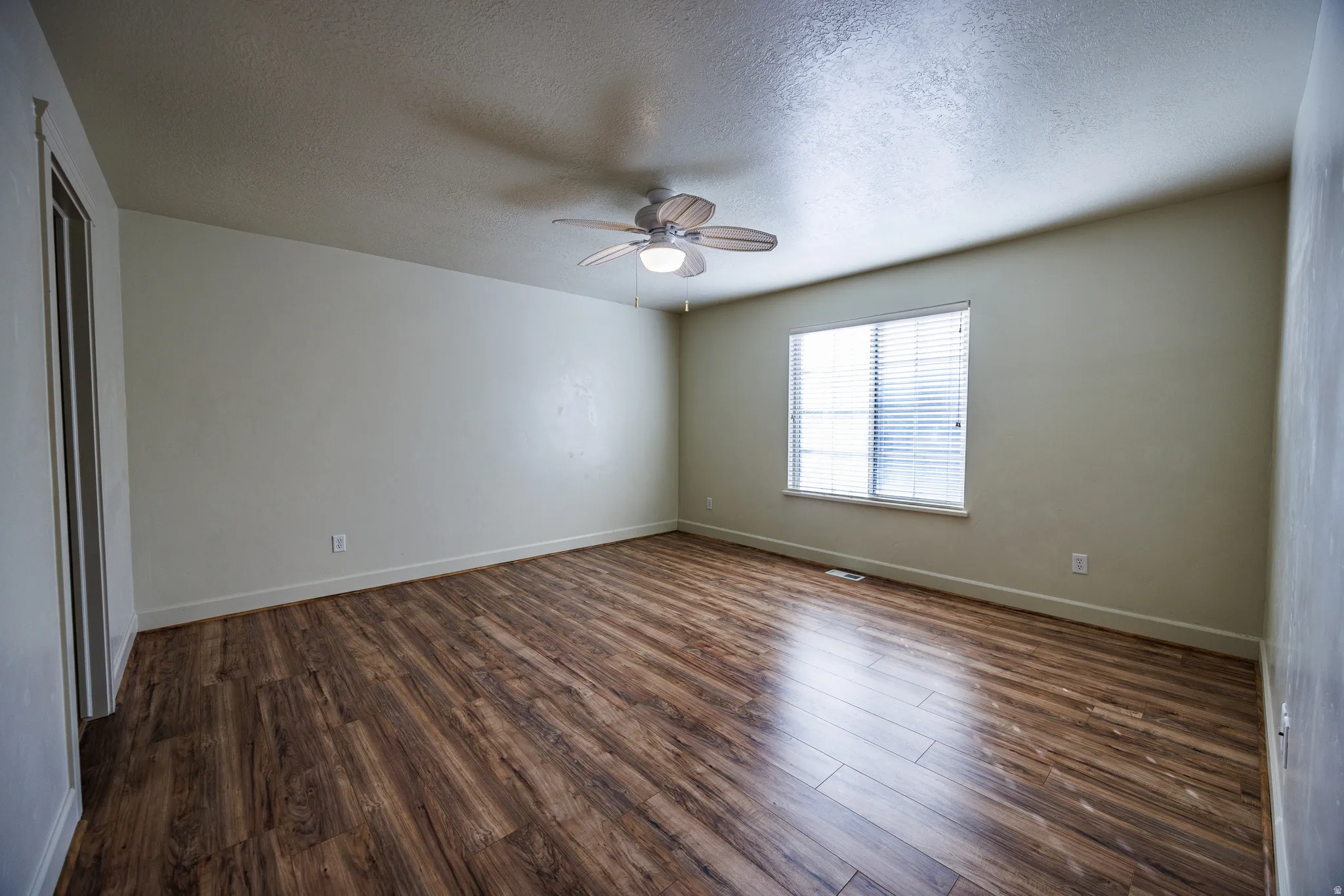 Spacious room featuring wood-finish flooring, a ceiling fan with integrated lighting, a large window with blinds, and neutral wall tones