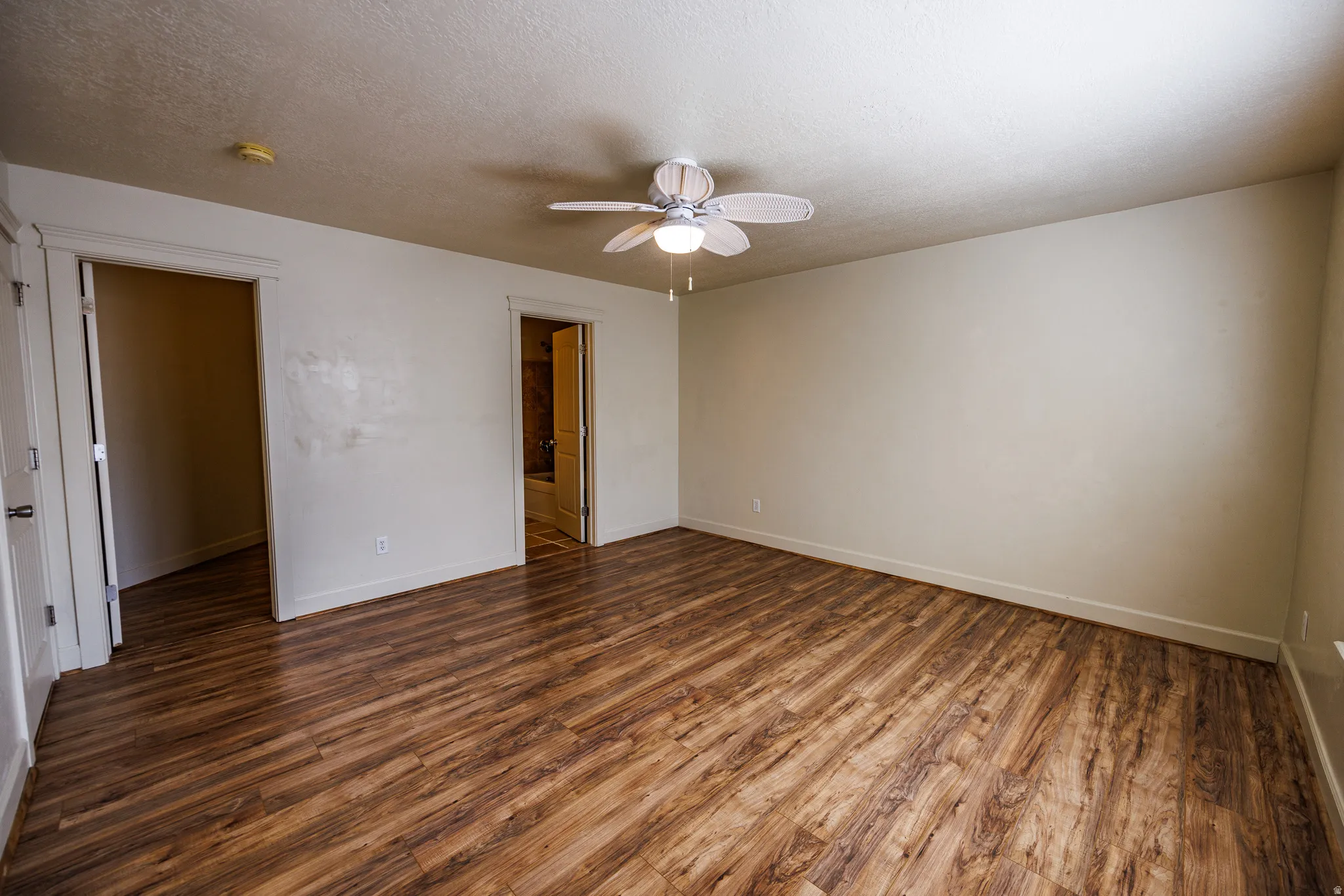 Expansive room featuring wood-finish flooring, a ceiling fan with integrated lighting, two doorways, and neutral-toned walls