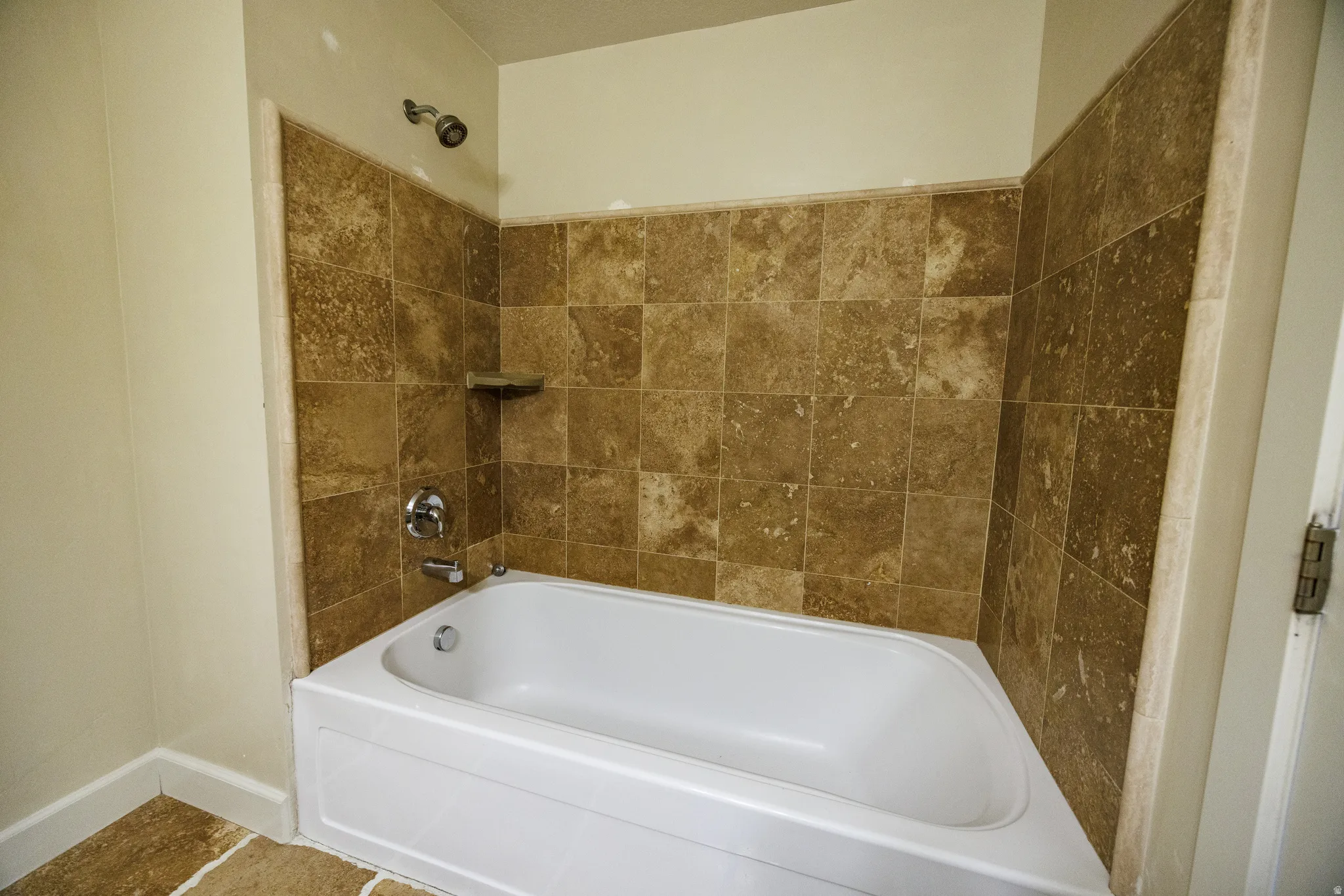 White bathtub with chrome fixtures, surrounded by earth-toned square wall tiles and a matching corner shelf