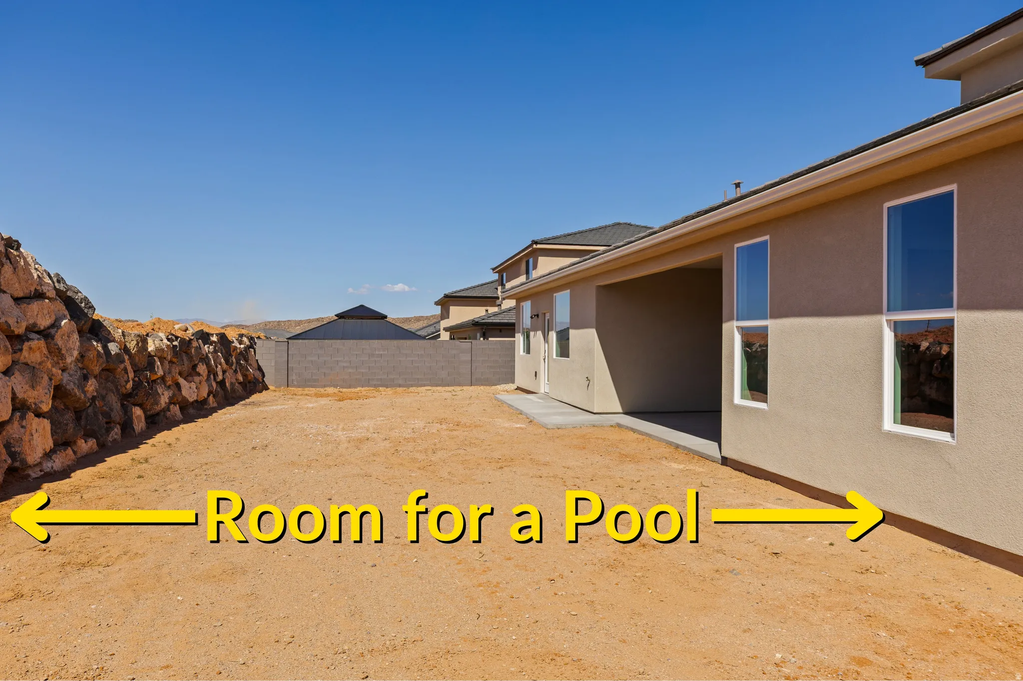 Spacious backyard featuring a stucco exterior, multiple windows, a covered patio, and a stone retaining wall