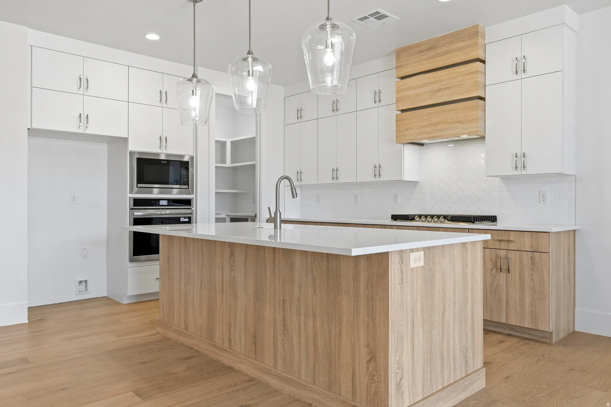 Modern kitchen featuring a central island with a light wood-finish base, three pendant lights, white countertops, and a stainless steel faucet