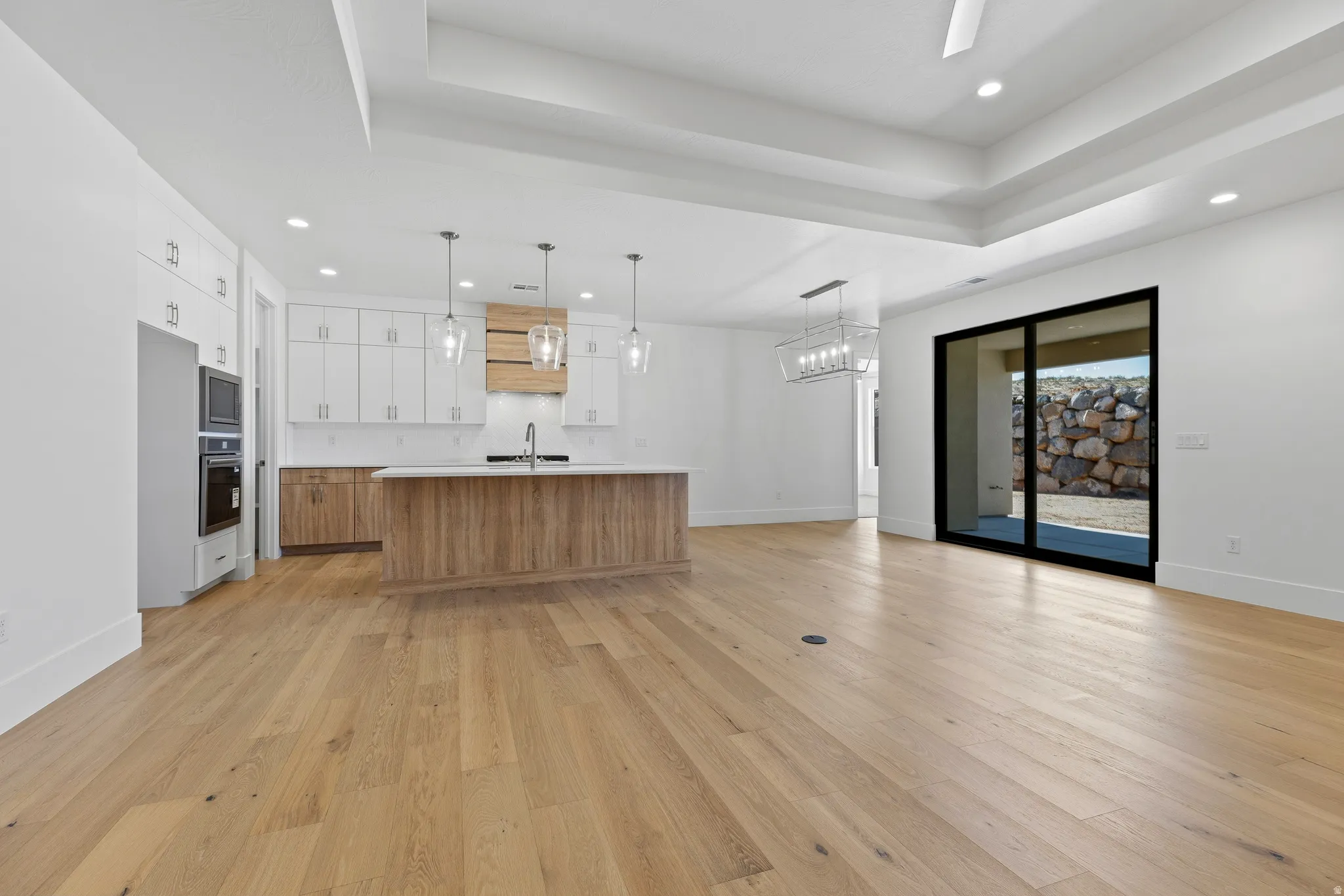 Open-concept living area featuring light wood-finish flooring, a contemporary kitchen island with a light countertop, integrated stainless steel appliances, a tray ceiling, and a large black-framed sliding glass door