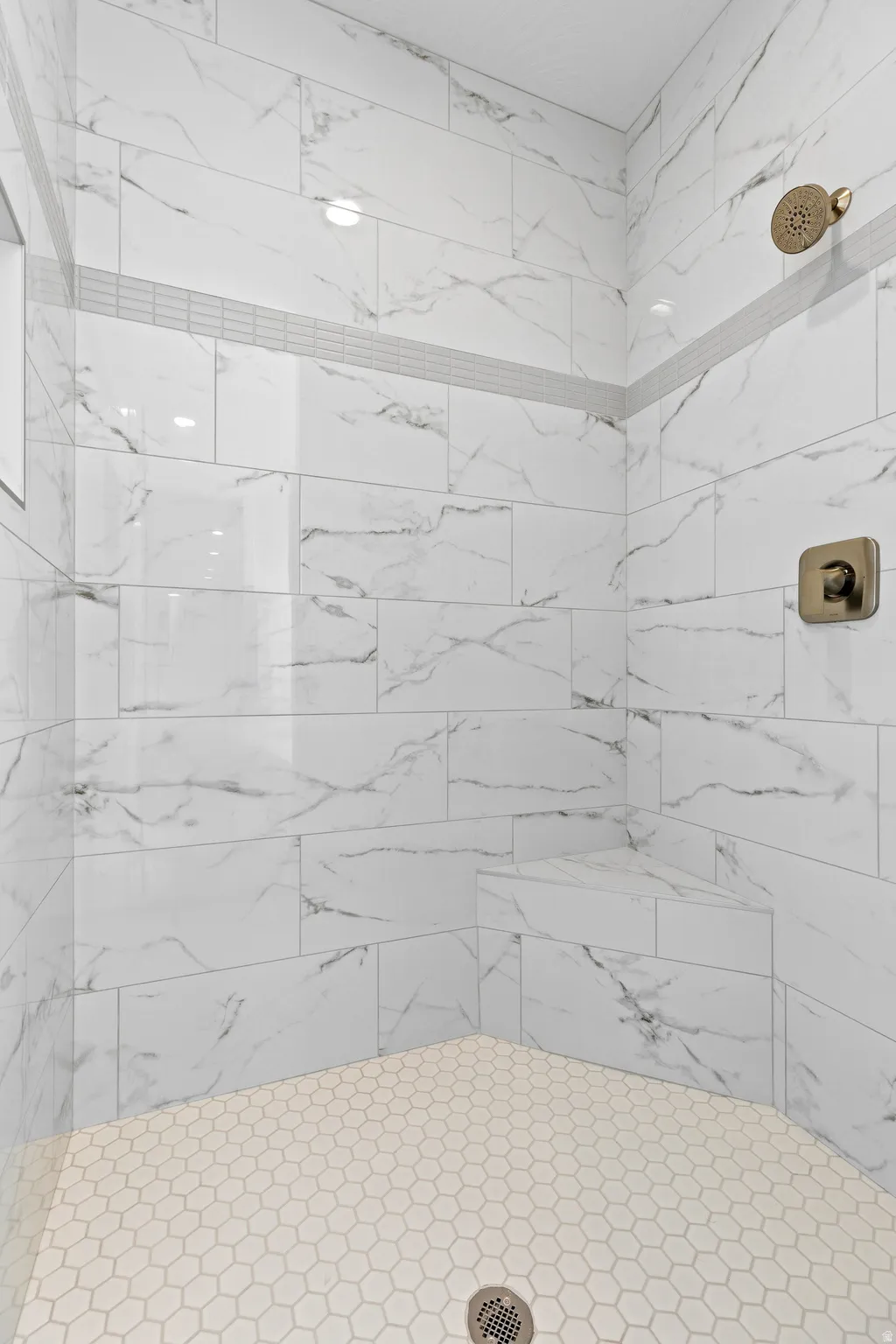 Walk-in shower featuring large format white tile walls with gray veining, a decorative mosaic accent band, a built-in shower bench, a hexagonal tile floor, and a bronze shower head and control