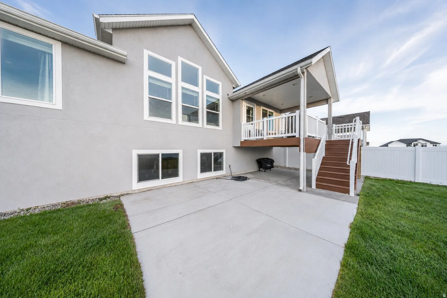 Exterior stucco finish with multiple large windows, a ground-level concrete patio, a covered upper deck with white railings, a brown wood-finish staircase, and a white privacy fence