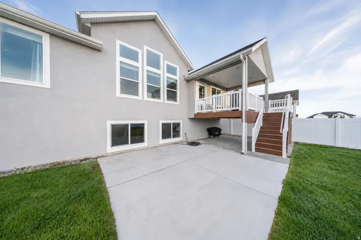 Exterior stucco finish with multiple large windows, a ground-level concrete patio, a covered upper deck with white railings, a brown wood-finish staircase, and a white privacy fence