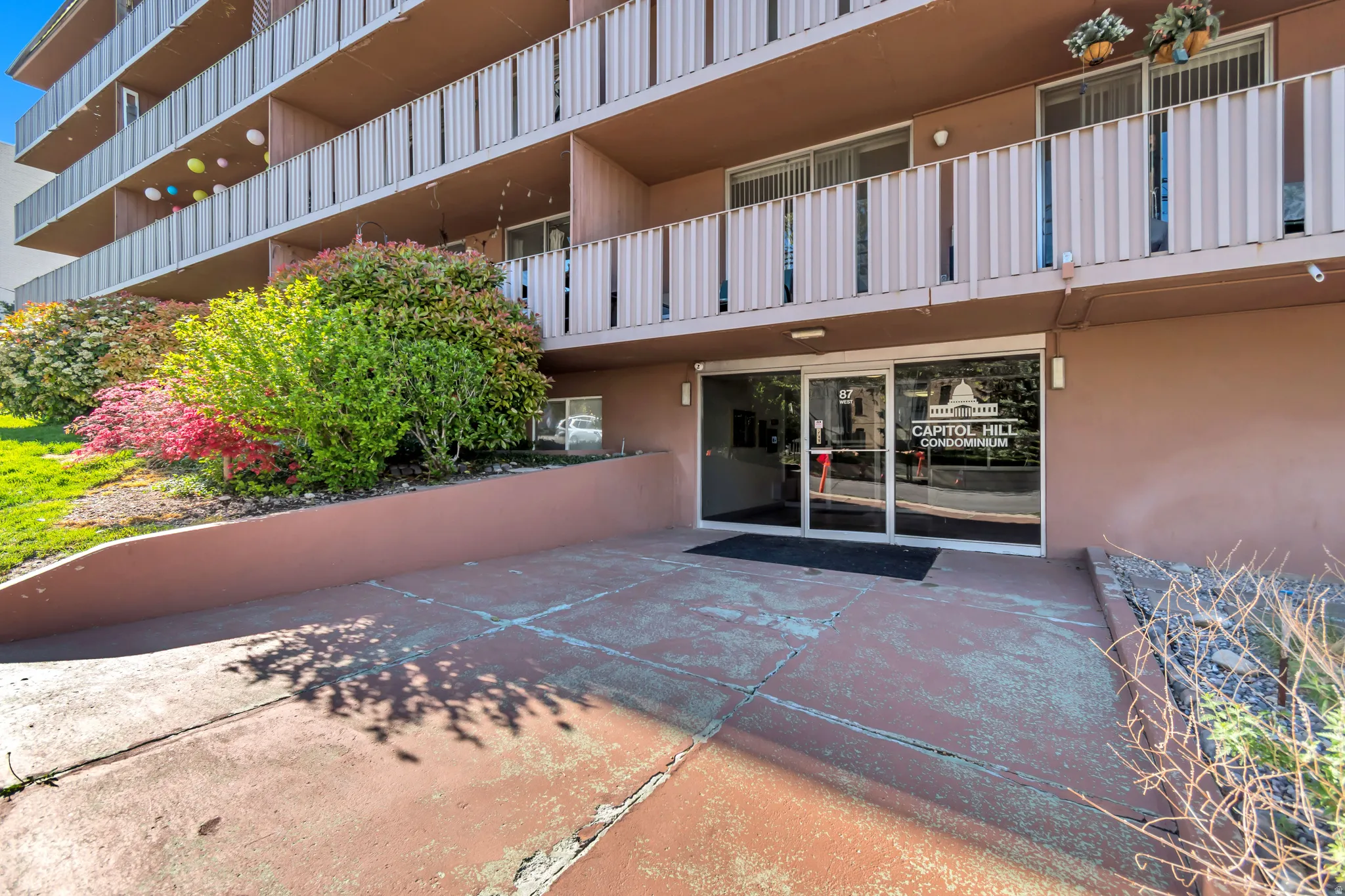 Capitol Hill Condominium building entrance featuring a glass door, a dark entryway mat, and an adjacent landscaped area with mature plantings