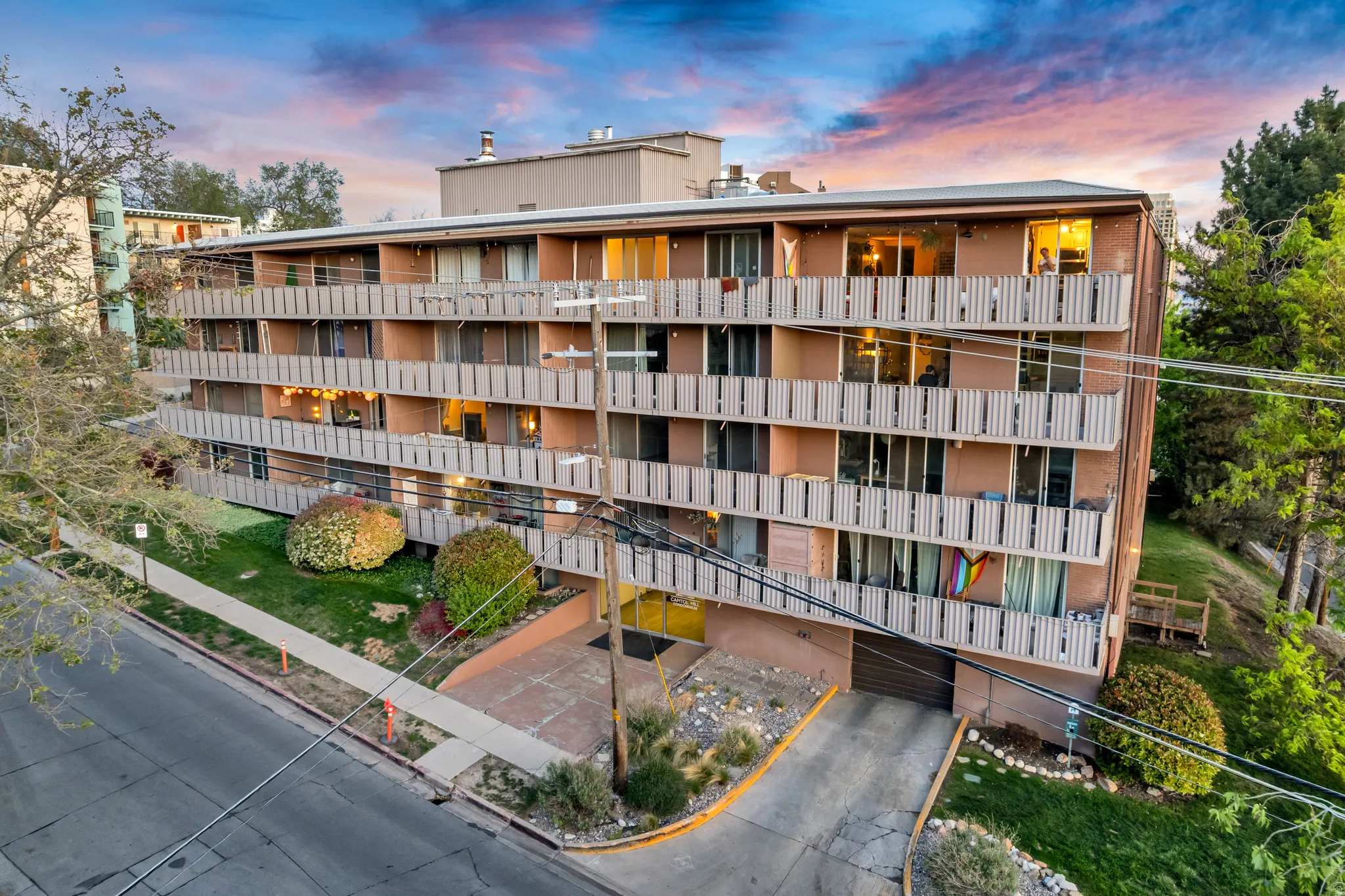 Multi-story residential building featuring private balconies, a paved drive-up entry, mature landscaping, and a street-level sidewalk
