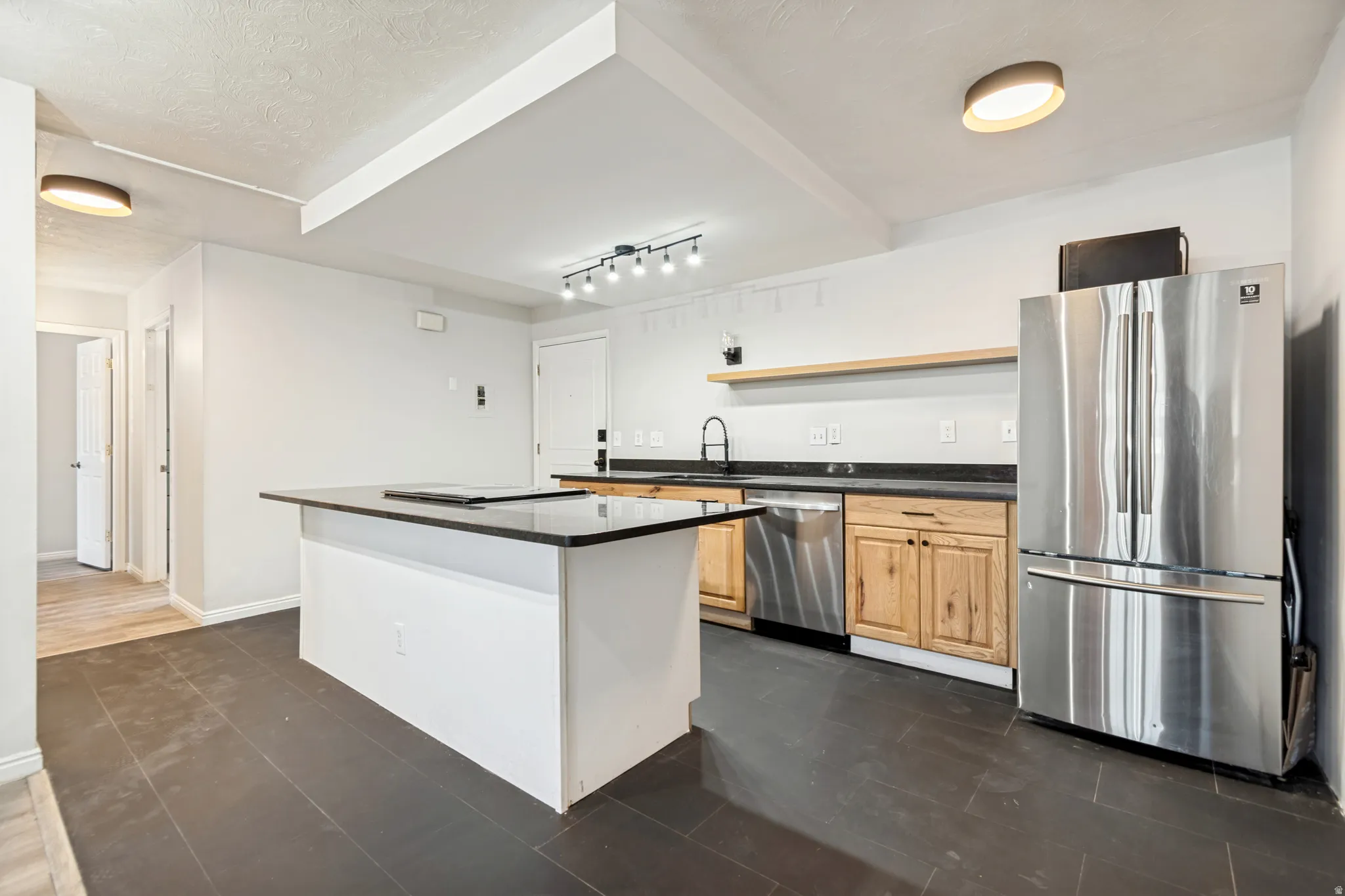 Open-concept kitchen featuring dark tile flooring, a center island with a dark countertop, light wood cabinetry, stainless steel appliances, and a floating wood shelf
